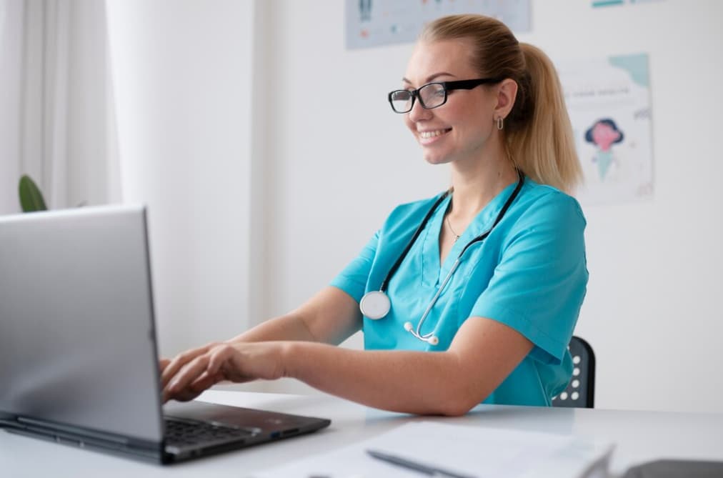 A smiling nurse with glasses typing on a laptop at a desk